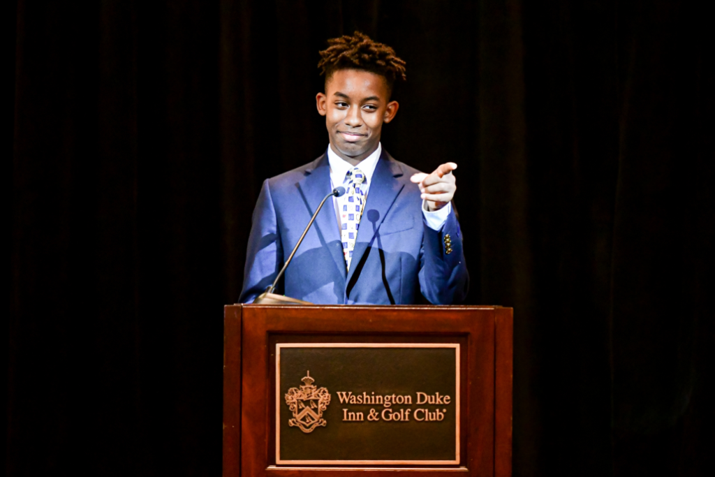 A young man presents behind a podium and points to the audience