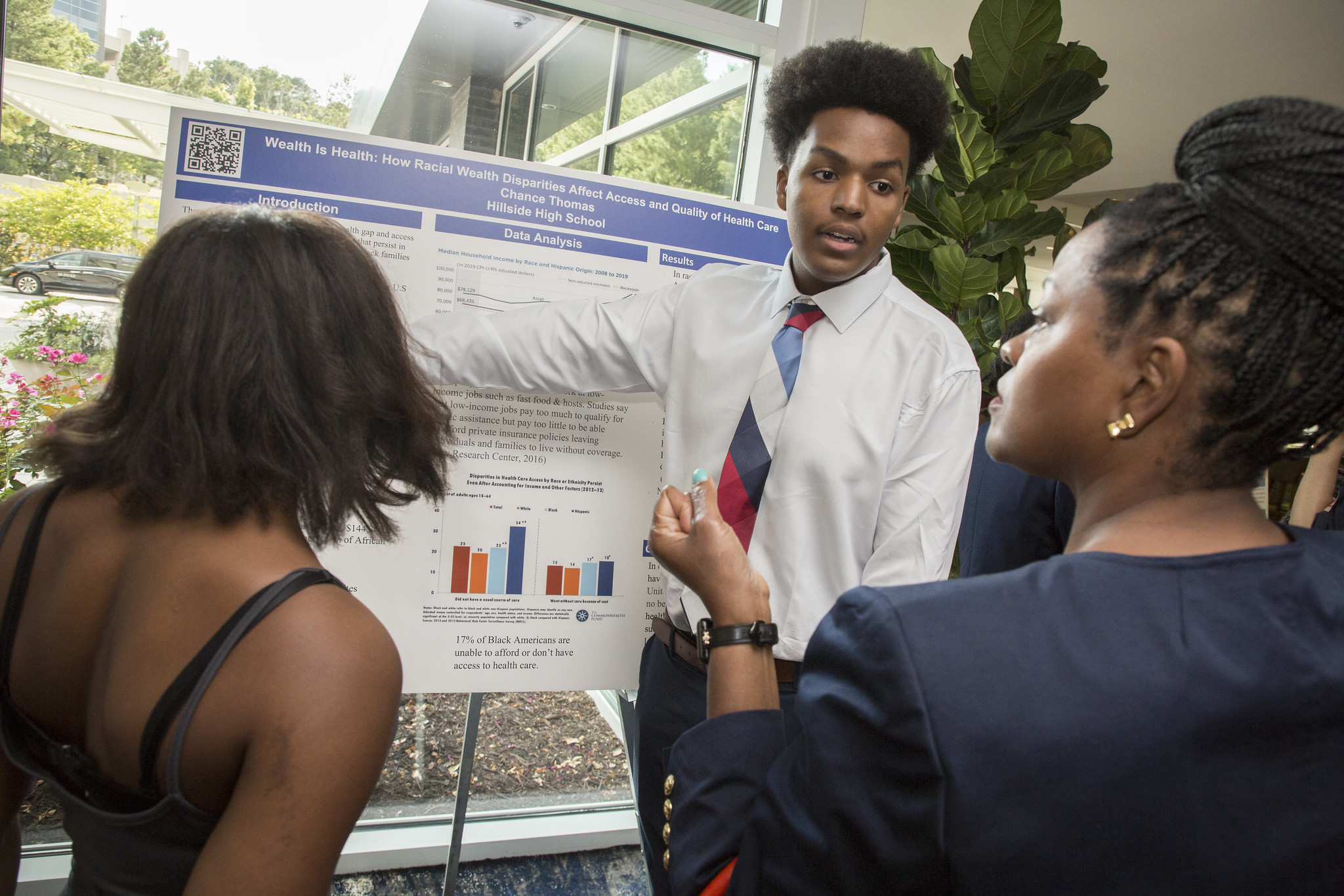 A young man participating in the Young Scholars program presents his research on a poster board to two people